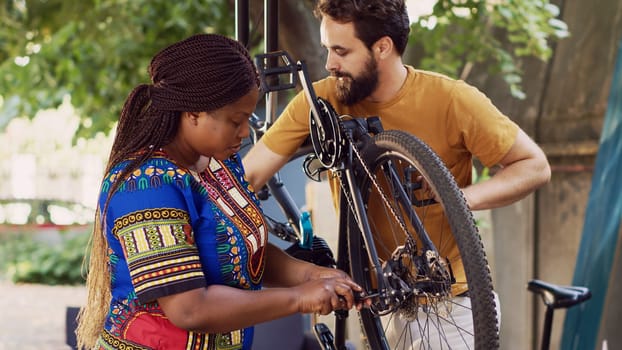 Multiethnic couple working together to repair and maintain damaged bike for summer cycling. Caucasian boyfriend helps girlfriend in choosing suitable tool for fixing bicycle hub and axle in yard.