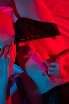 A faceless woman lies in a bedroom in a blue-red neon light and uses a pink vibrator. Close-up of the abdomen
