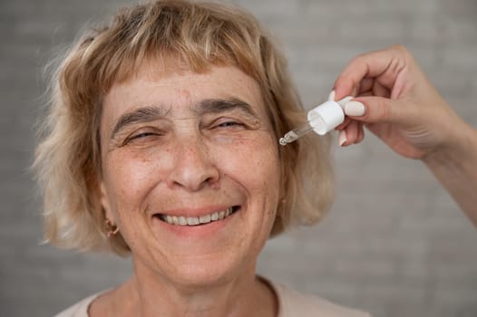 Close-up portrait of an old woman applying hyaluronic acid serum with a pipette. Anti-aging face care
