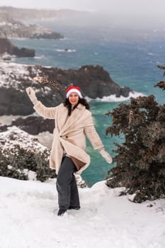 Outdoor winter portrait of happy smiling woman, light faux fur coat holding heart sparkler, posing against sea and snow background.