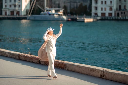 Happy blonde woman in a white suit and hat posing at the camera against the backdrop of the sea.