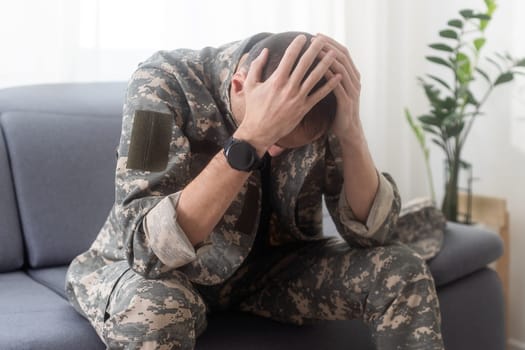 Portrait of calm serious Caucasian military man wearing camouflage uniform and cap sitting on a sofa, having depressed facial expression. High quality photo