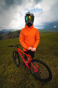 Wide angle vertical frame top angle. Portrait of a bearded mountain biker with his bicycle against a background of mountains and clouds in the summer surrounded by green grass.