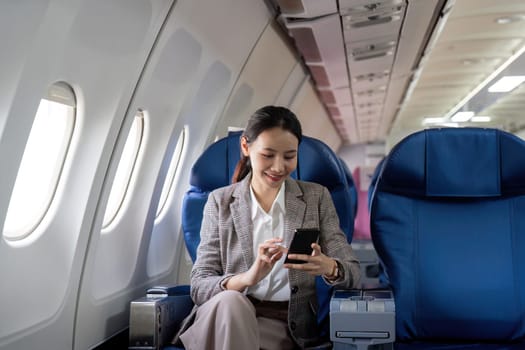 Asian young businesswoman successful or female entrepreneur in formal suit in a plane sit in a business class seat and uses a smartphone during flight. Traveling and Business concept.