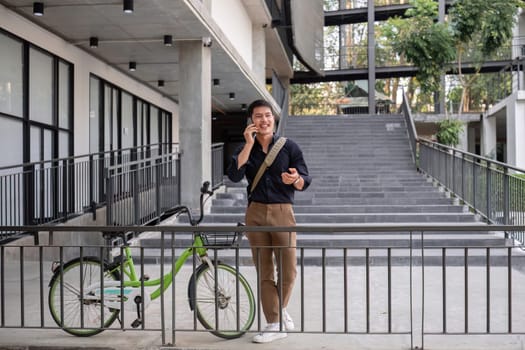 A young Asian businessman rides a bicycle to work. Standing outside the office talking on the phone and talking about business. Concept of reducing energy and reducing air pollution..