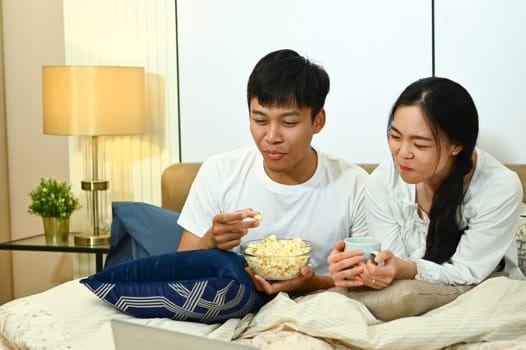 Happy marriage couple watching TV and eating popcorn on couch at home.