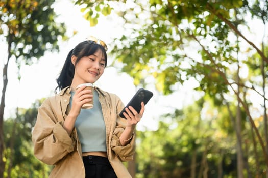 Smiling young lady sitting in the park drinking coffee and checking her mobile phone.