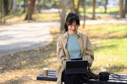 Charming young female freelancer sitting on park bench and working on digital tablet.