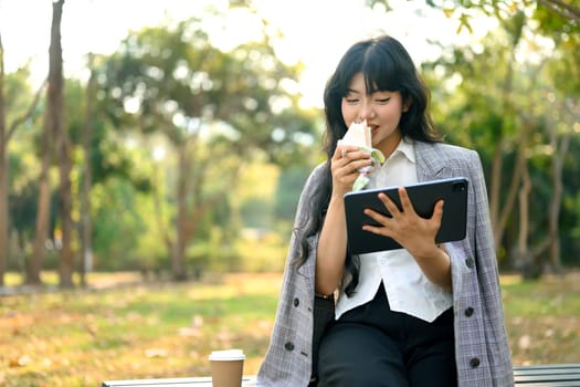 Beautiful young businesswoman with digital tablet having lunch on bench in park.