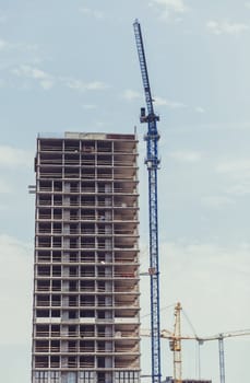 three building cranes at the background of a multi-storey building under construction. New houses will wait for families