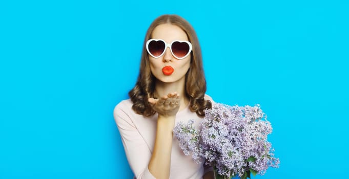 Portrait of beautiful happy young woman blowing kiss with bouquet of fresh lilac flowers in heart shaped sunglasses on blue studio background