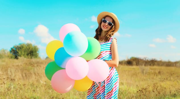 Happy smiling young woman with bunch of colorful balloons in summer straw hat on field on blue sky background