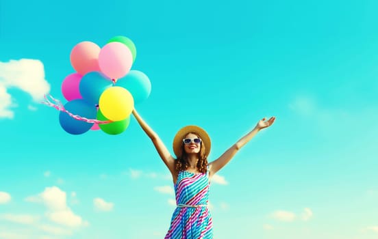 Happy joyful young woman with bunch of colorful balloons in summer straw hat having fun on blue sky background