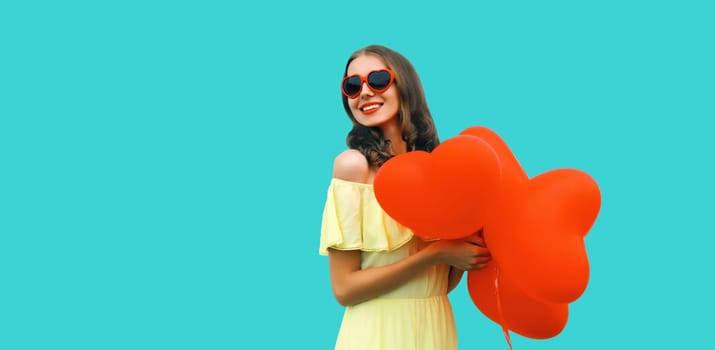 Cute portrait of happy smiling young woman with red heart shaped balloon in sunglasses on blue studio background