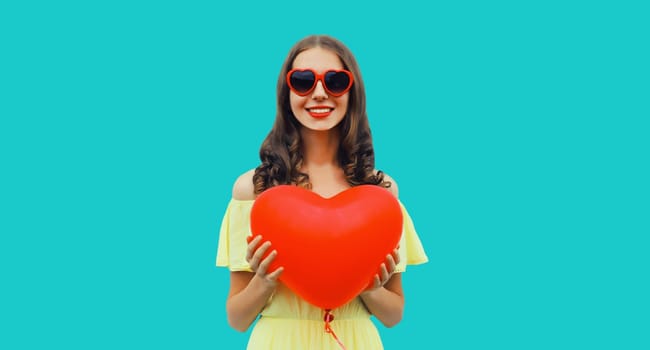 Cute portrait of happy smiling young woman with red heart shaped balloon in sunglasses on blue studio background