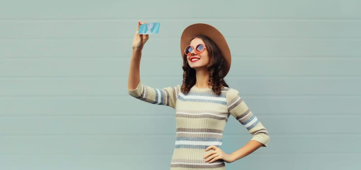 Portrait of stylish happy smiling young woman taking selfie with mobile phone in round hat, sunglasses on gray background