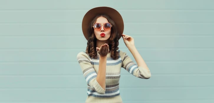 Portrait of stylish beautiful young woman blowing kiss in round hat, sunglasses posing on gray background