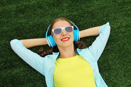 Portrait of happy smiling young woman listening to music in headphones while lying on grass in summer park