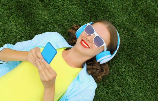 Happy young woman holding phone listening to music in headphones while lying on grass in summer park