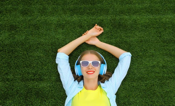 Portrait of happy smiling young woman listening to music in headphones while lying on grass in summer park