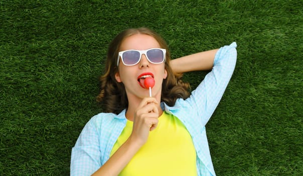 Portrait of happy teenager girl resting with lollipop lying on green grass in summer park, top view