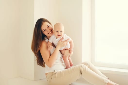 Happy smiling young mother playing with cute baby sitting together in white room at home