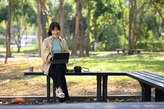 Full body of delighted young woman using digital tablet sitting on bench in park.