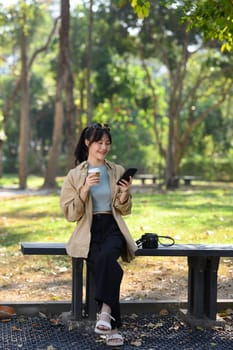 Full length portrait of young woman drinking coffee and using smartphone on bench in park.