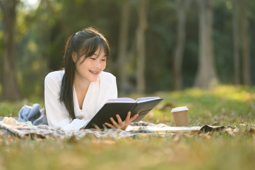 Peaceful young Asian woman lying on picnic blanket and reading her reading her favorite book.