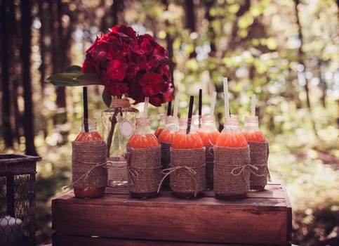 bottles of juice and red flowers on the rustic table. Vintage summer wedding table decoration.