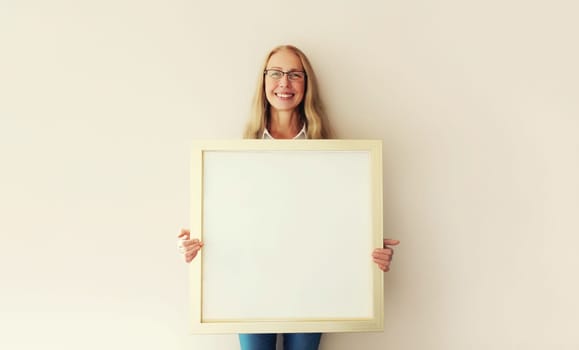 Happy smiling mature woman decorating interior, hanging white blank photo frame, wooden picture, artwork mockup on wall in new house