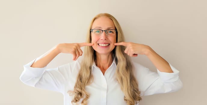 Portrait of happy smiling middle-aged woman points with fingers to her white clean teeth in eyeglasses on white studio background