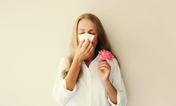 Sick exhausted woman suffering from pollen holds flower sneezing blow nose using tissue on white studio background