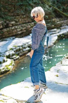 beautiful sexy girl in nature among a mountain river and a rocky canyon in a denim jumpsuit smiles cheerfully and enjoys life on vacation in the summer