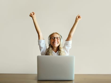 Happy joyful middle aged woman listening to music in headphones working with laptop computer sitting at desk at home