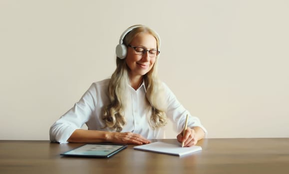 Happy relaxing middle aged woman listening to music in headphones working with digital tablet computer sitting at desk at home