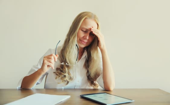 Tired overworked middle-aged woman employee suffering from headache, rubbing her dry eyes, working with tablet computer sitting at desk in office