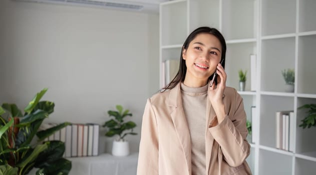 Asian businesswoman talking on the phone in an online business meeting in a modern home office decorated with lush green plants..