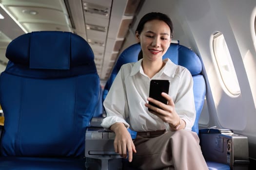 Young Asian woman checks business news on mobile phone, sitting near window in first class on airplane during flight, travel and business concept.