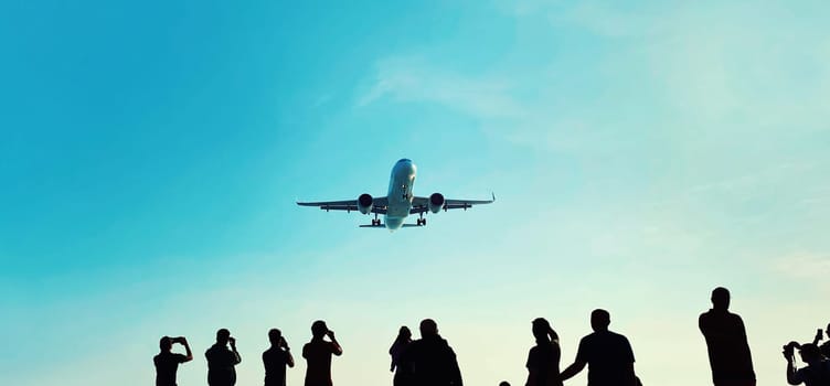 Silhouette of group of unknown people tourists take pictures a landing passenger airplane flying in blue sky background