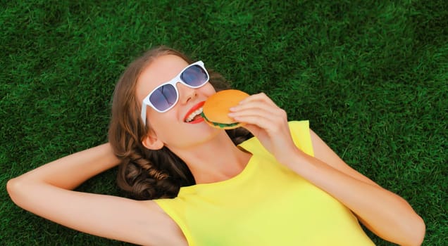 Portrait of happy smiling young woman with burger fast food lying on green grass in summer park
