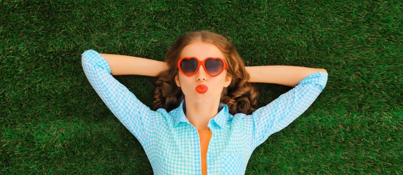 Summer portrait of happy young relaxing woman in red heart shaped sunglasses blowing kiss lying on green grass in the park