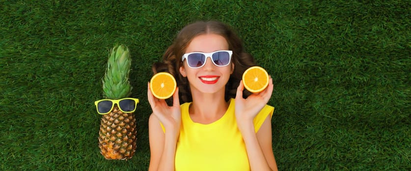 Summer portrait of happy smiling young relaxing woman with fruits, pineapple in sunglasses lying on green grass in the park