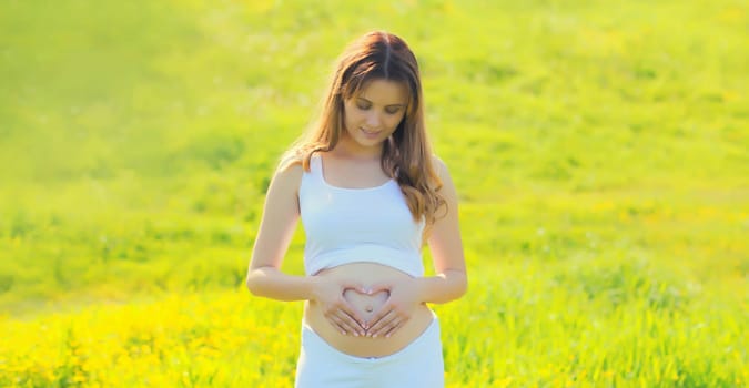 Happy young pregnant woman makes heart sign on her belly with her hands in summer park