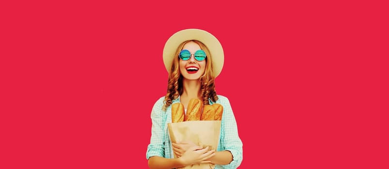 Portrait of happy cheerful smiling young woman holding grocery shopping paper bag with fresh long white bread baguette on red studio background