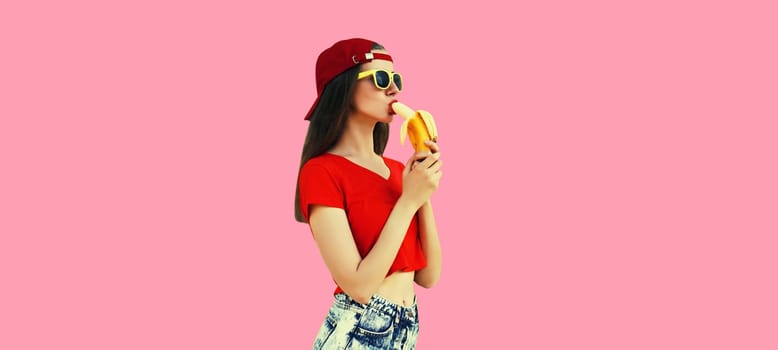 Stylish caucasian young woman eating banana in red baseball cap on pink studio background