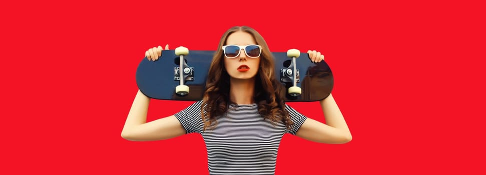 Portrait of stylish young woman posing with skateboard on red studio background
