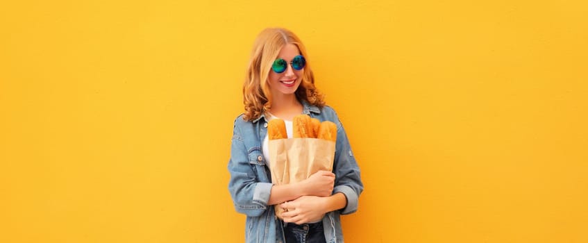Portrait of happy cheerful smiling young woman holding grocery shopping paper bag with fresh long white bread baguette on yellow studio background