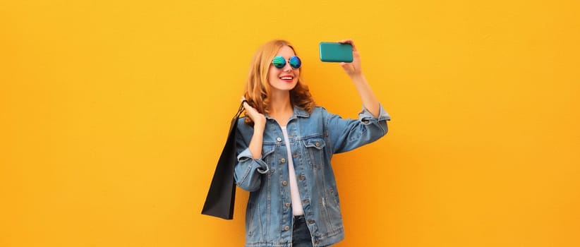 Portrait of beautiful happy smiling young woman taking selfie with phone holding black shopping bags on yellow studio background