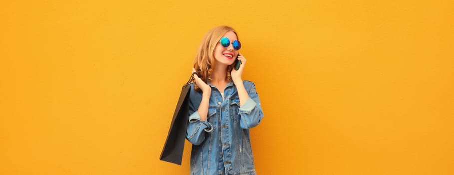 Beautiful happy young woman calling on smartphone with black shopping bag looking away in denim jacket on yellow studio background